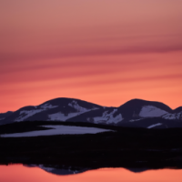 En realistisk bild av ett bergslandskap vid solnedgången, med orange och rosa himmel, snötäckta bergstoppar och en spegelblank sjö i förgrunden. Sigma 85mm f/8