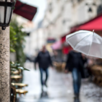 En realistisk bild av en stadsgata i Paris på en regnig dag, med våta kullerstenar, människor med paraplyer och en caféterrass i bakgrunden. Sigma 85mm f/1.4